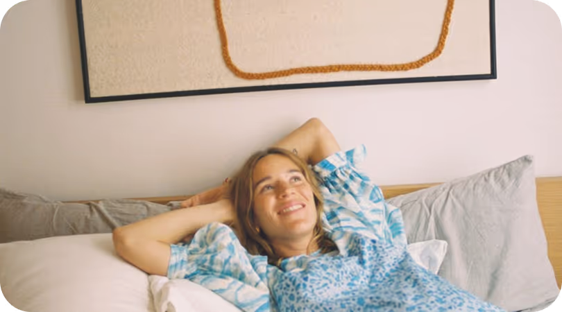 Jeune femme souriante allongée sur un lit avec des coussins et un tableau abstrait au mur.