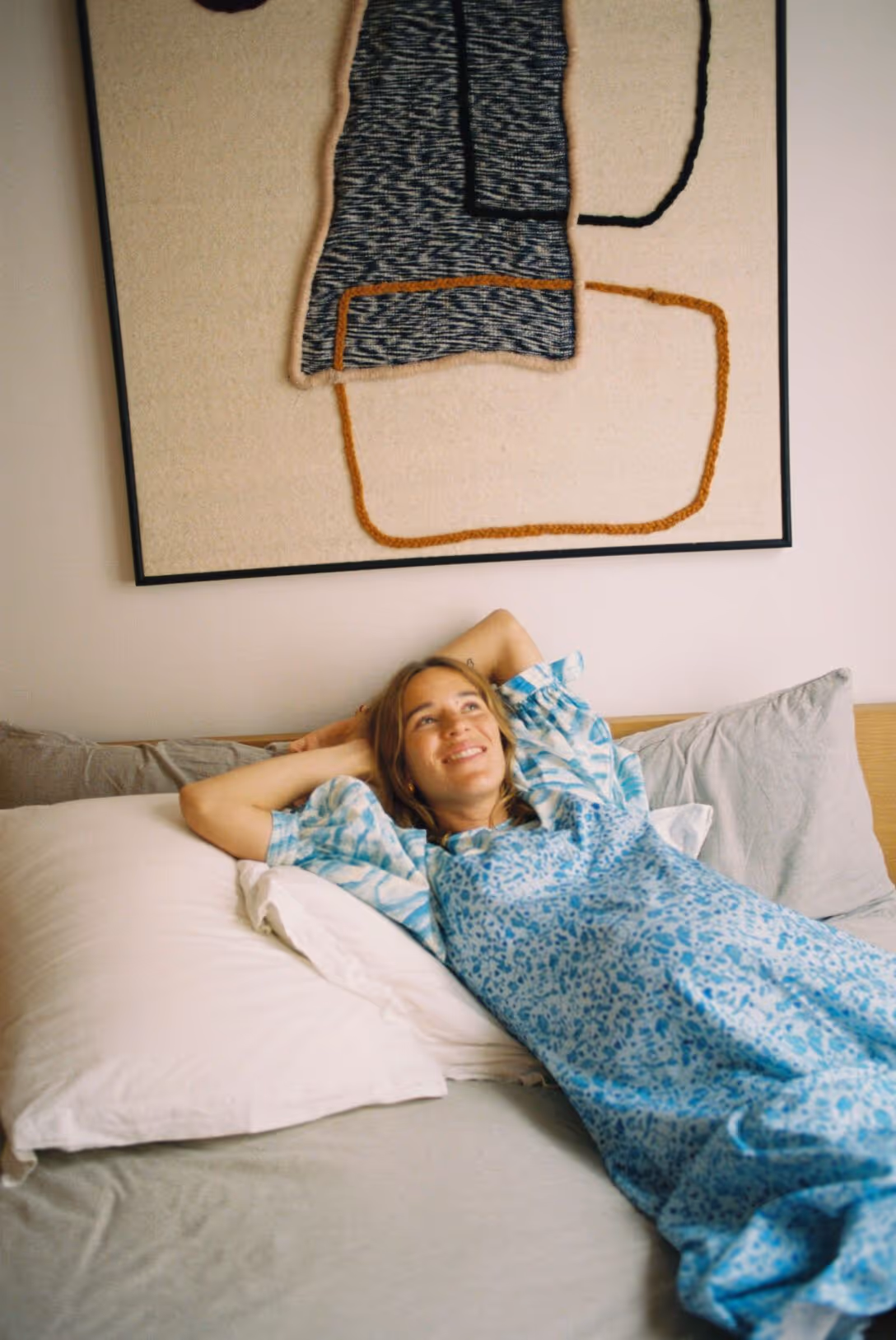 Femme souriante allongée sur un lit avec oreillers blancs et tableau abstrait au mur.
