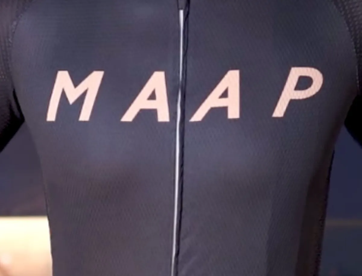 Closeup front view of a counterfeit MAAP cycling jersey showing oversized pink logo lettering on a dark navy fabric with a center zipper, worn on a cyclist indoors