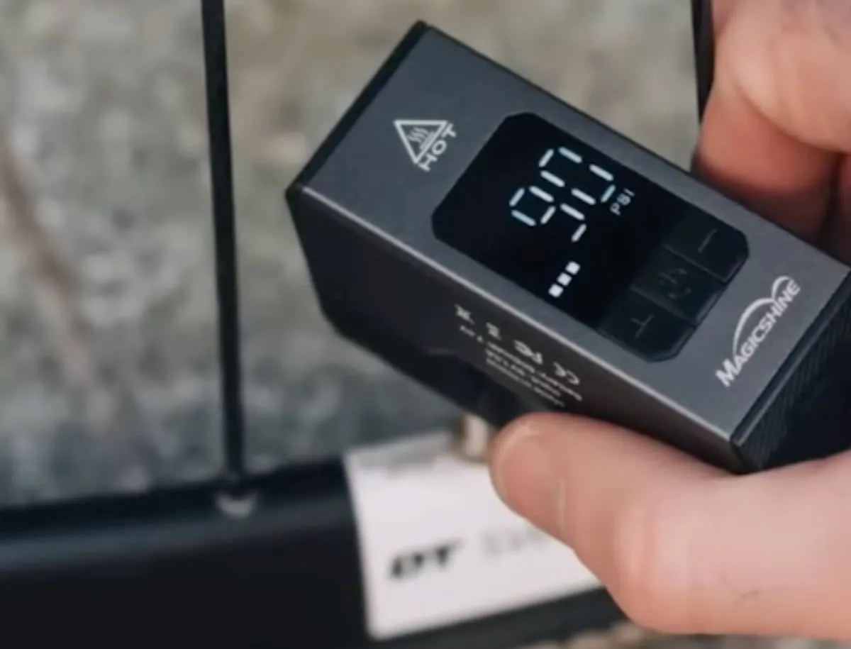 Hand holding a compact Magicshine AIRRO Mini portable electric tire inflator with digital PSI display screen showing current tire pressure, connected to a road bike tire valve outdoors