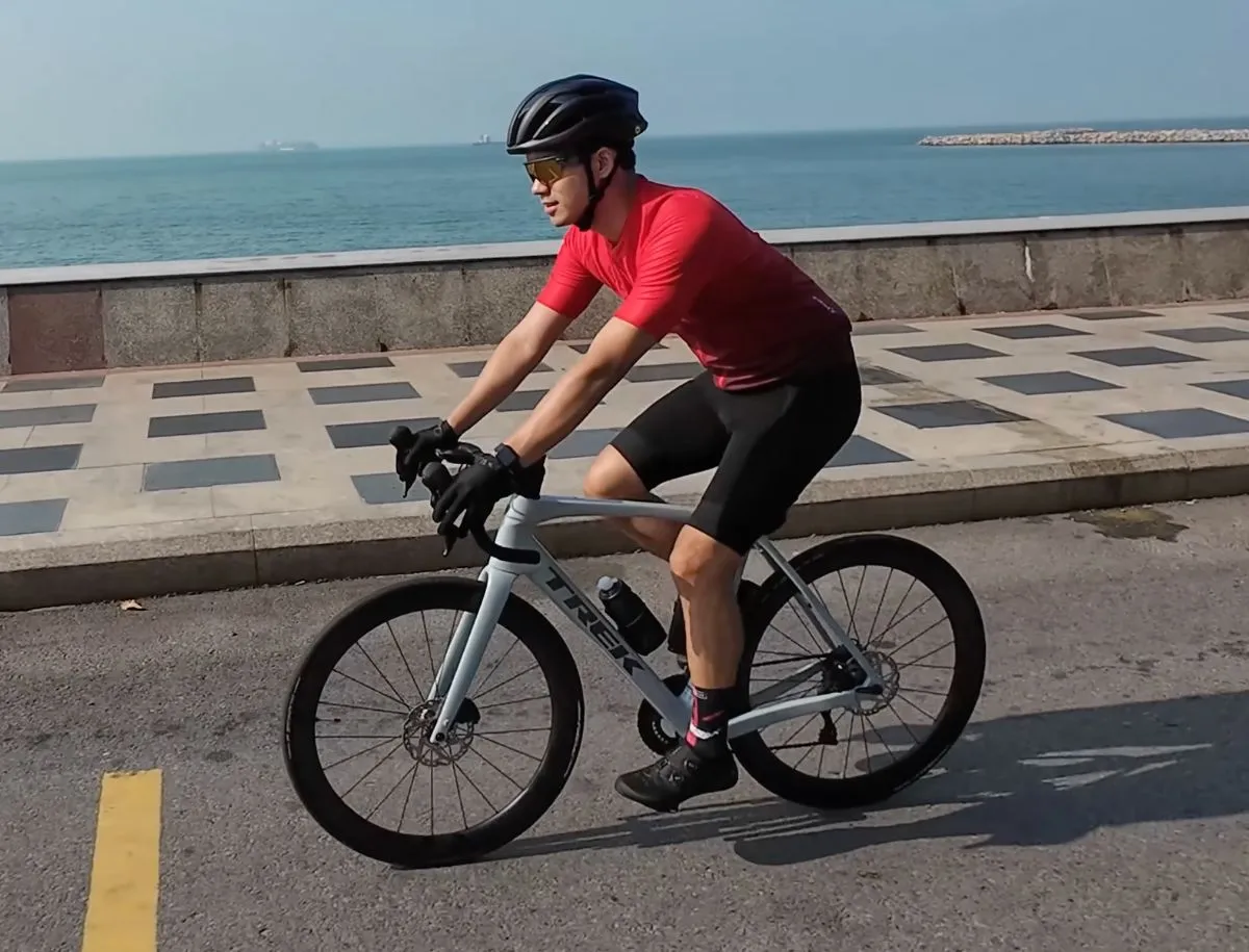 Cyclist wearing red jersey, black bib shorts, helmet, and Oakley sunglasses riding a Trek road bike along a coastal road