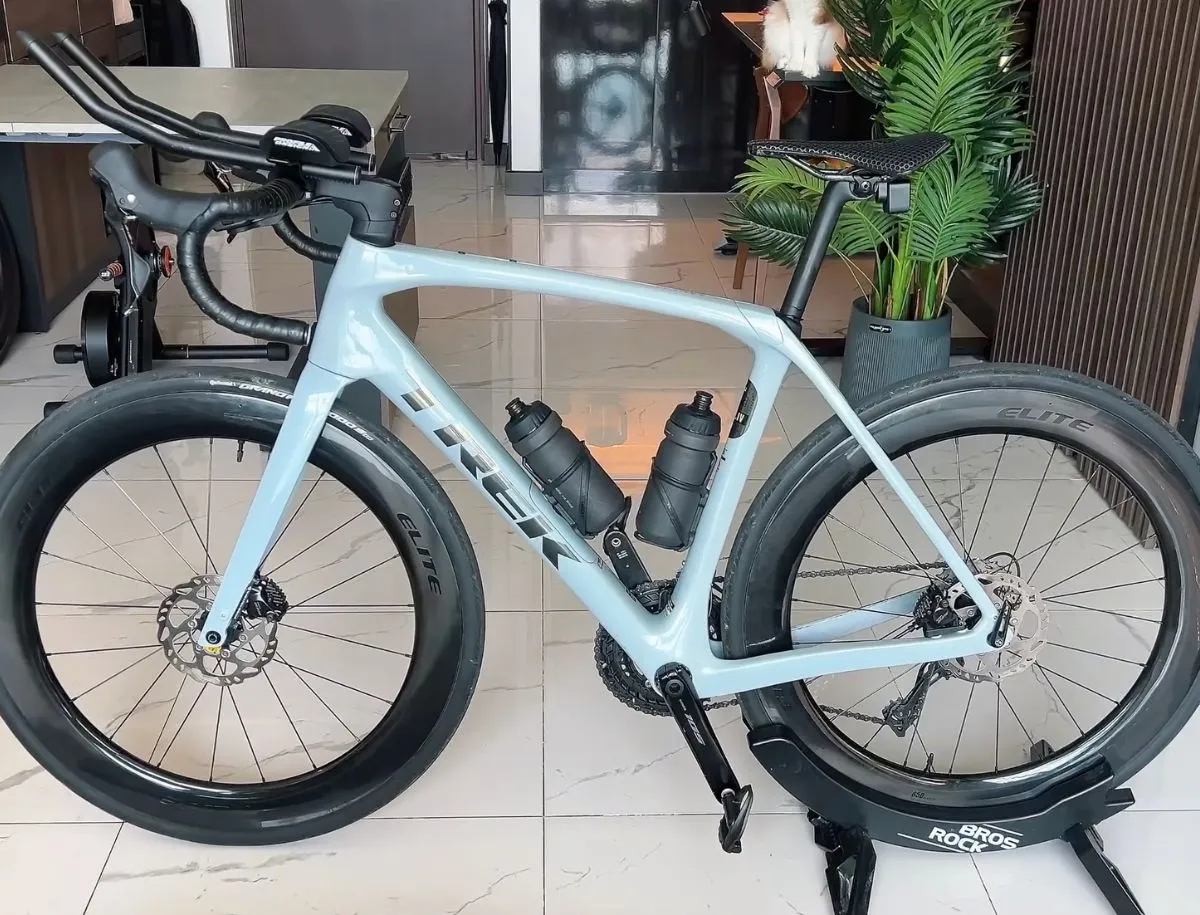 Light blue Trek carbon road bike with triathlon cockpit, deep-section Elite carbon wheels, dual bottle cages, and disc brakes, displayed on a Bros Rock bike stand indoors