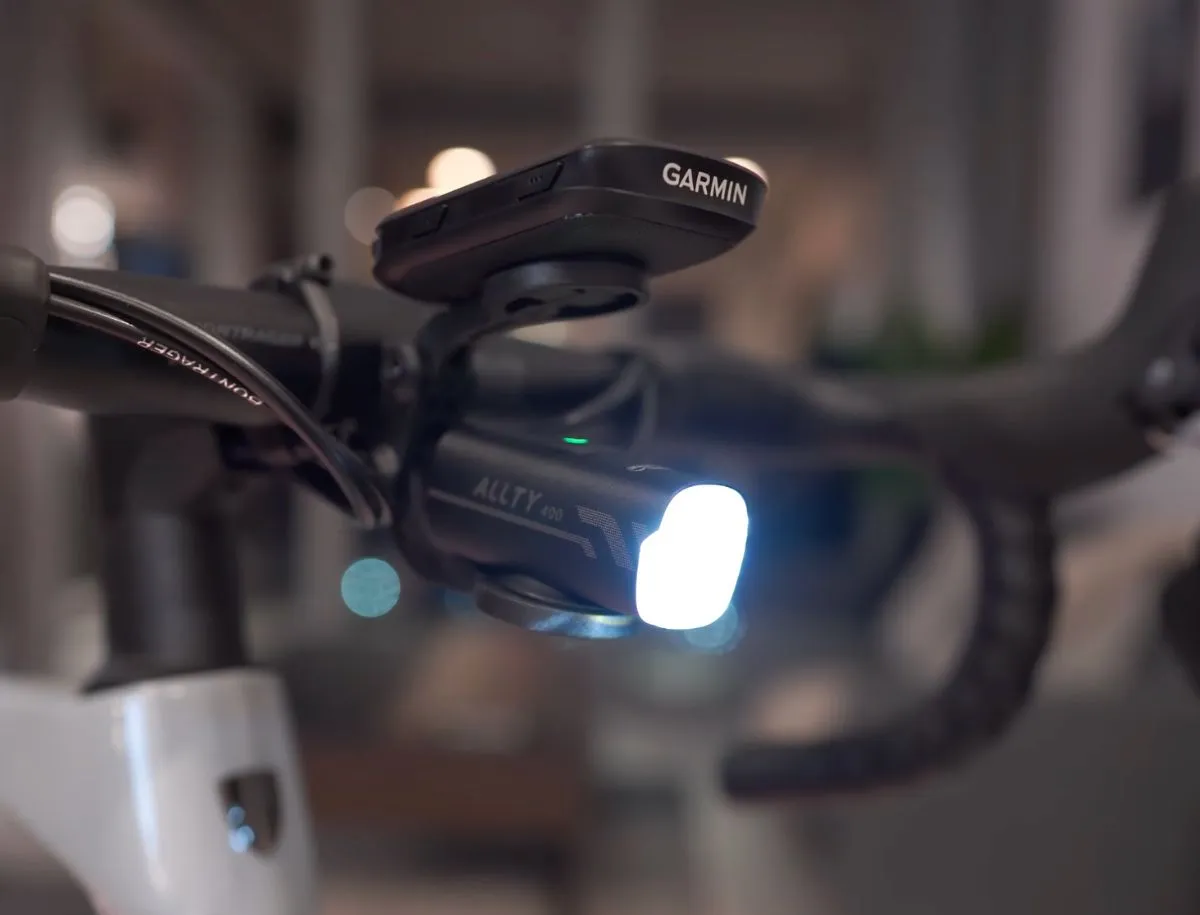 Magicshine Allty 400 front bike light mounted on road bike handlebars beneath a Garmin cycling computer, with the light powered on showing a bright white beam.