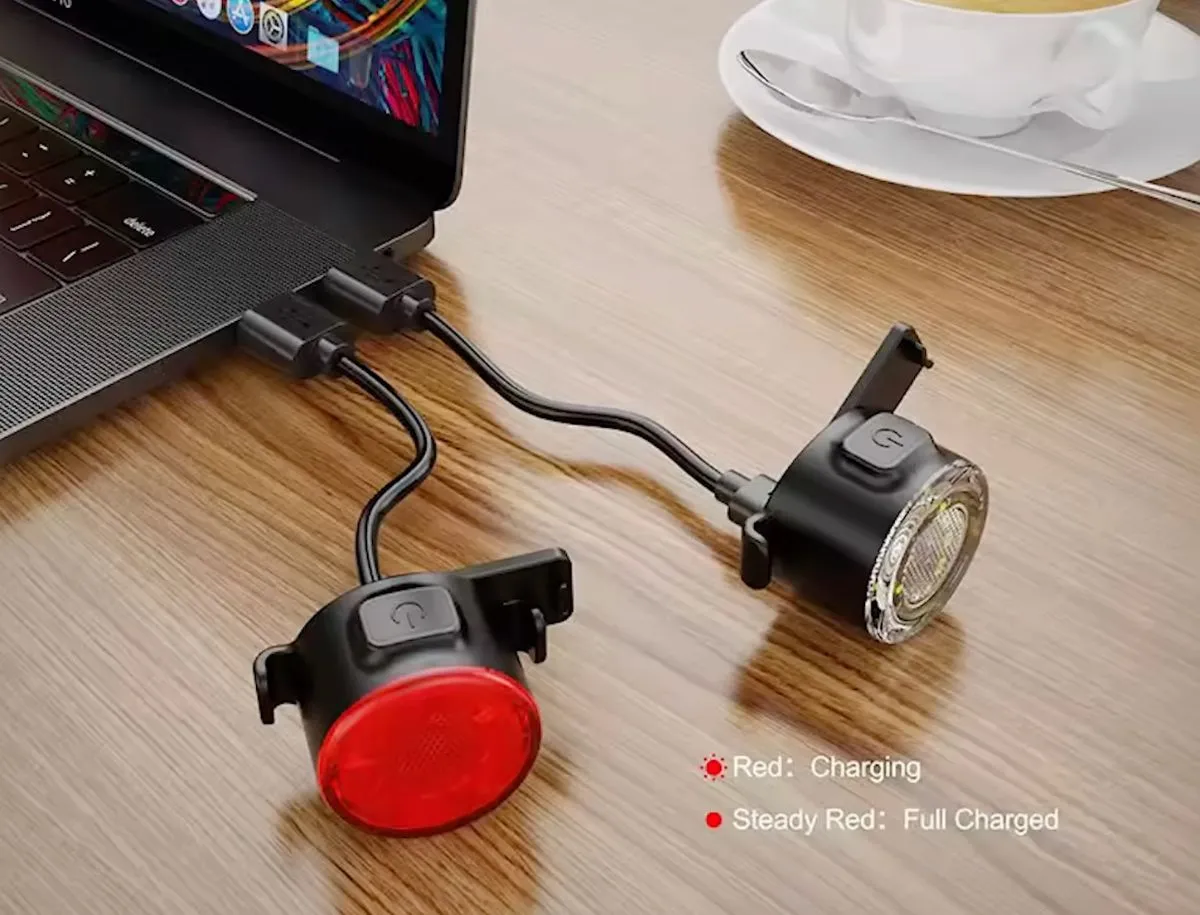 AliExpress front and rear bike light combo set charging via split USB cable connected to a MacBook, with charging indicator labels showing red for charging and steady red for full charge.