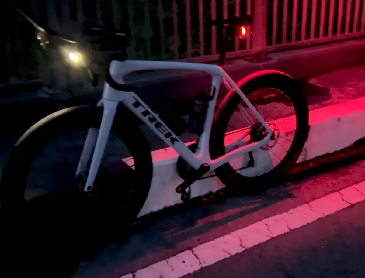 White Trek road bike parked on a dark urban street at night with a small red rear light glowing on the seatpost, red light washing across the road surface and white road markings.