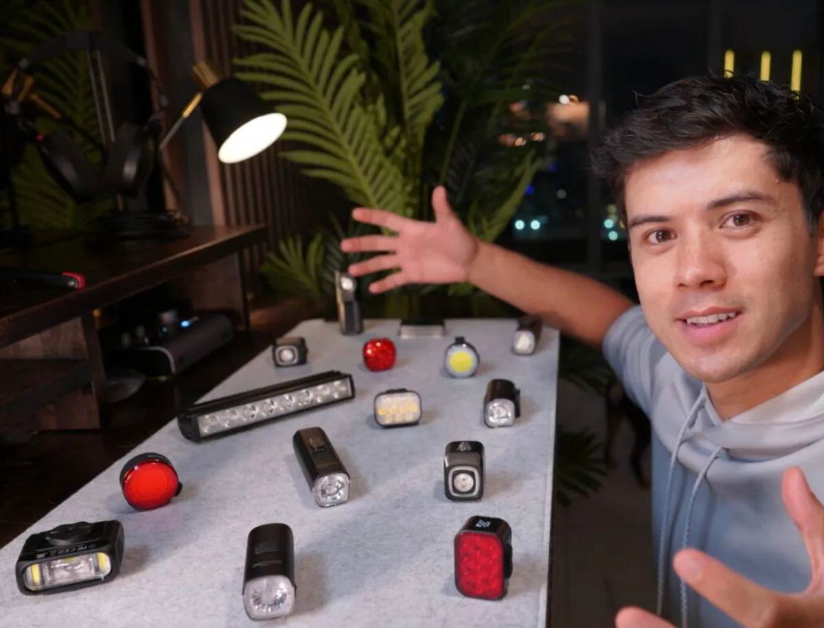 Cyclist Mike Dee presenting 12 AliExpress bike lights laid out on a table, including front lights, rear lights, and bar-mounted options of various sizes and styles
