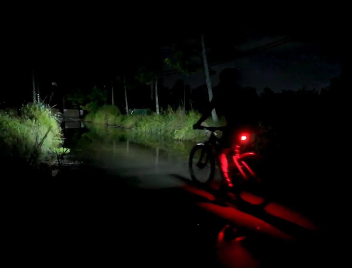  Cyclist riding at night on a dark rural road with a bright red rear bike light illuminating the road surface and surrounding vegetation behind them.
