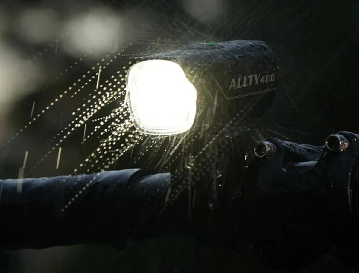 Magicshine Allty 400 front bike light mounted on handlebars glowing bright white in heavy rain, with water droplets visible across the lens and body.