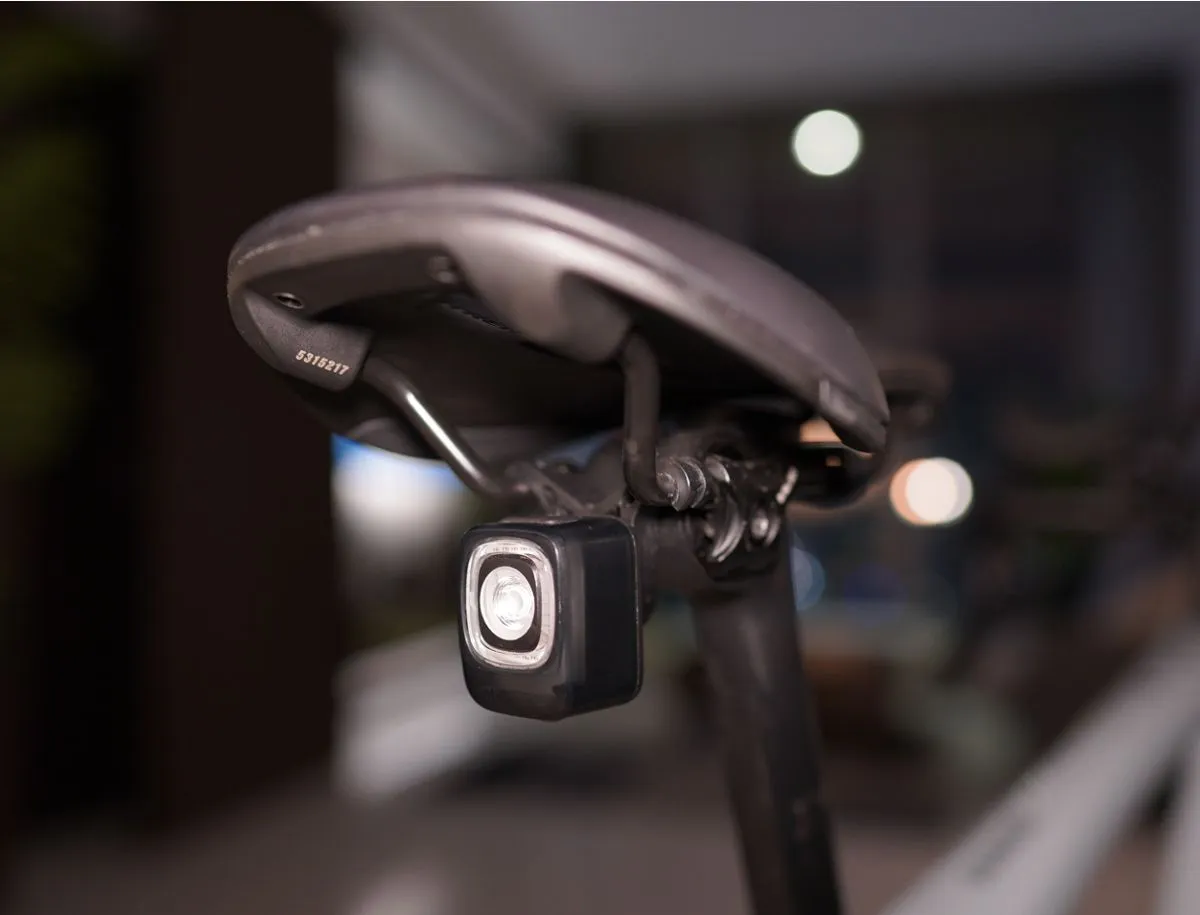 Magicshine SEEMEE 200 v3.0 rear bike light mounted under a road bike saddle on a seatpost, photographed at night with bokeh background lighting