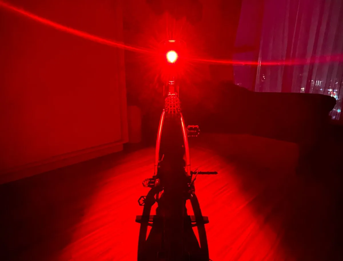 Magicshine SEEMEE 200 v3.0 rear bike light illuminated at full brightness inside a dark room, casting a wide 360-degree red light arc across the floor, walls, and bike frame to demonstrate road-surface visibility projection