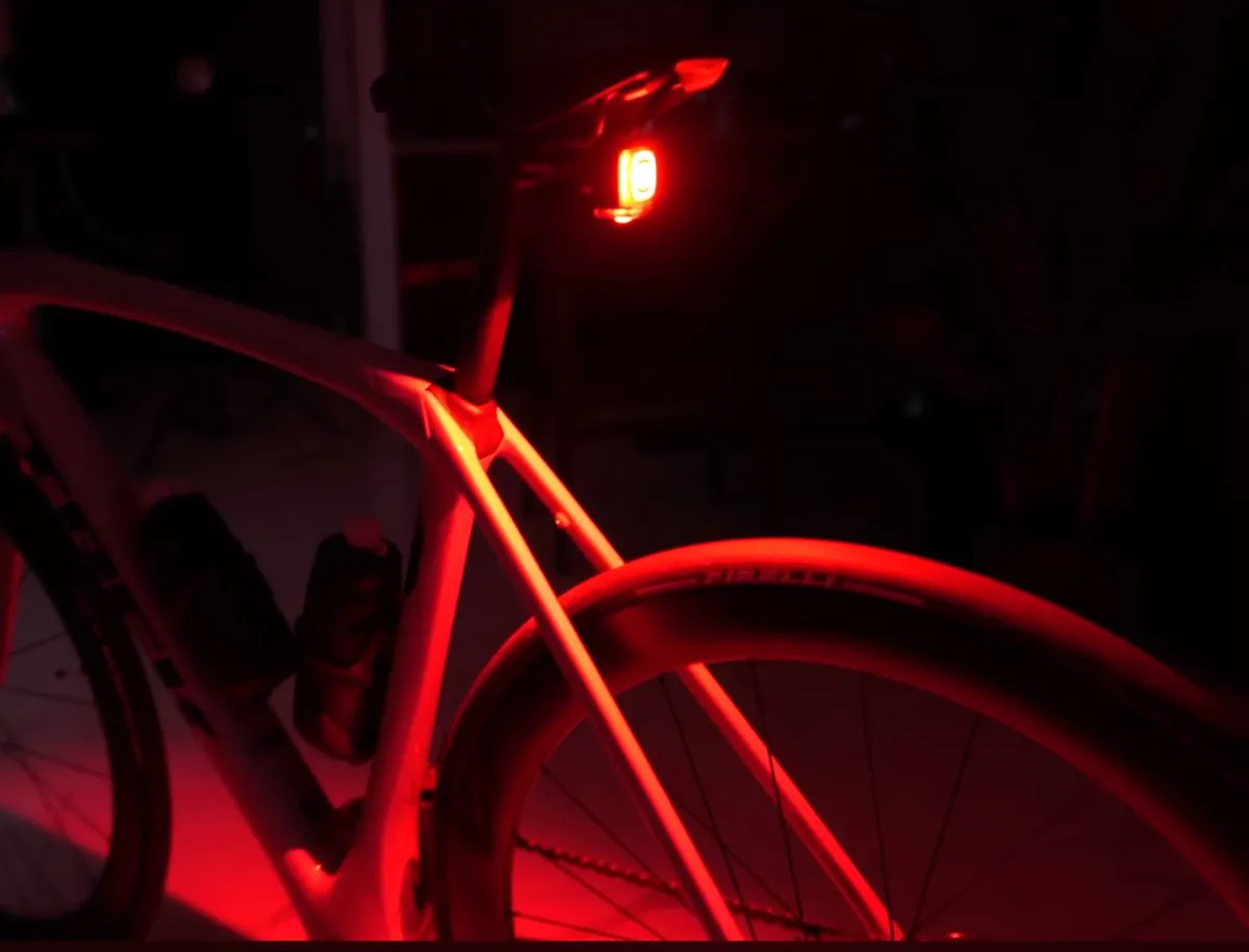 Magicshine Seemee 200 V3 rear bike light glowing bright red on the seatpost of a road bike at night, casting red light across the frame, rear wheel, and Pirelli tire in total darkness