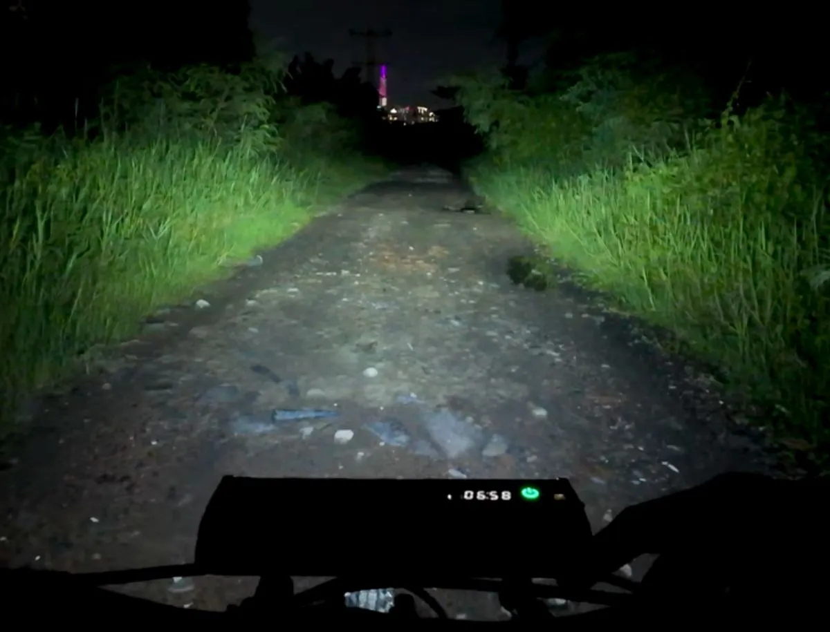 POV shot from a cyclist's perspective on a dark, rocky gravel path at night, lit by the NEWBOLER 10,000 lumen bike light mounted on the handlebars, showing wide 180-degree flood illumination across the trail edges and vegetation, with the LED display reading 6:58 remaining battery time