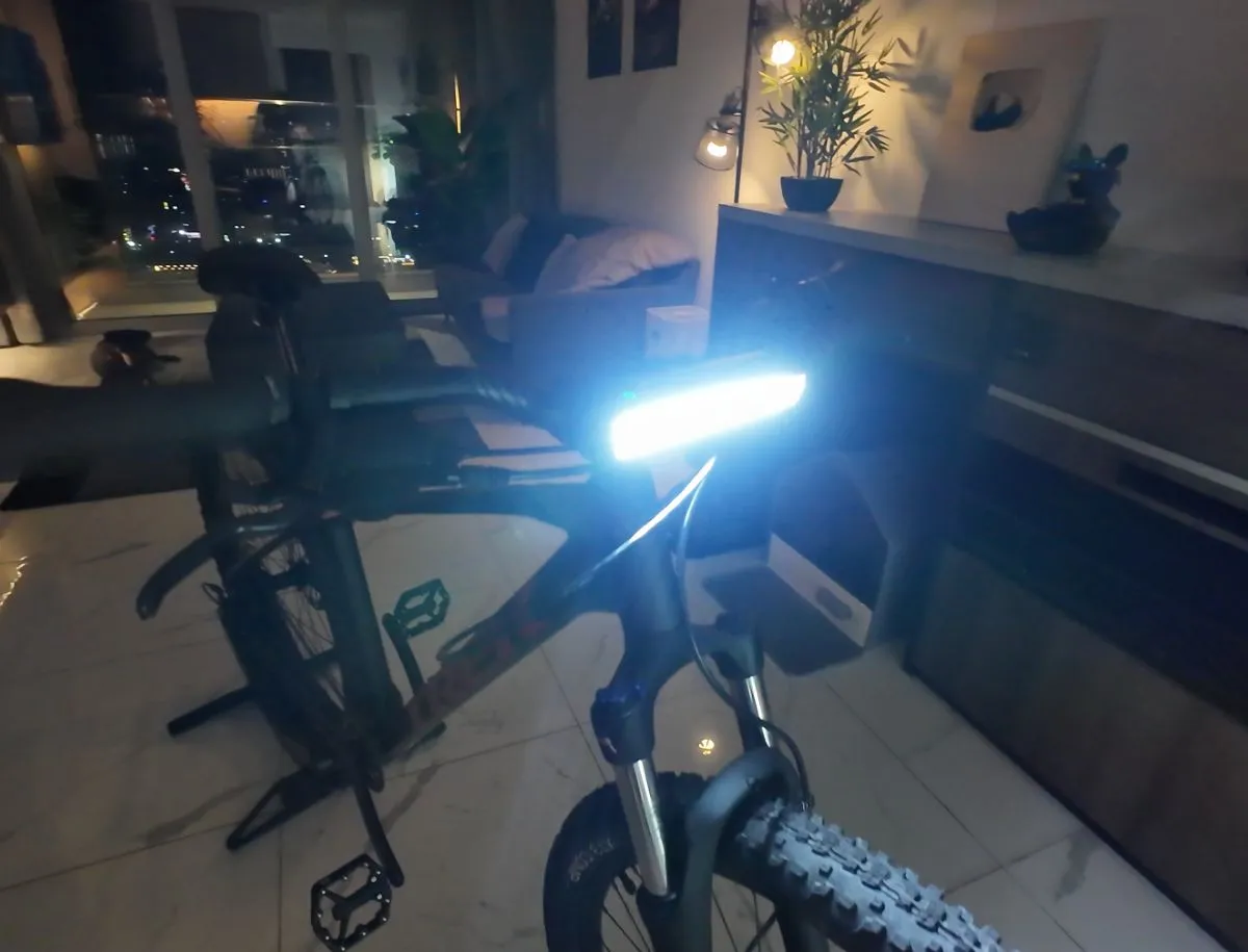 NEWBOLER 10,000 lumen bike light mounted on the handlebars of a mountain bike in an indoor setting at night, emitting a wide, intensely bright white beam, with a city skyline visible through the window in the background and a second bike on a trainer stand visible behind