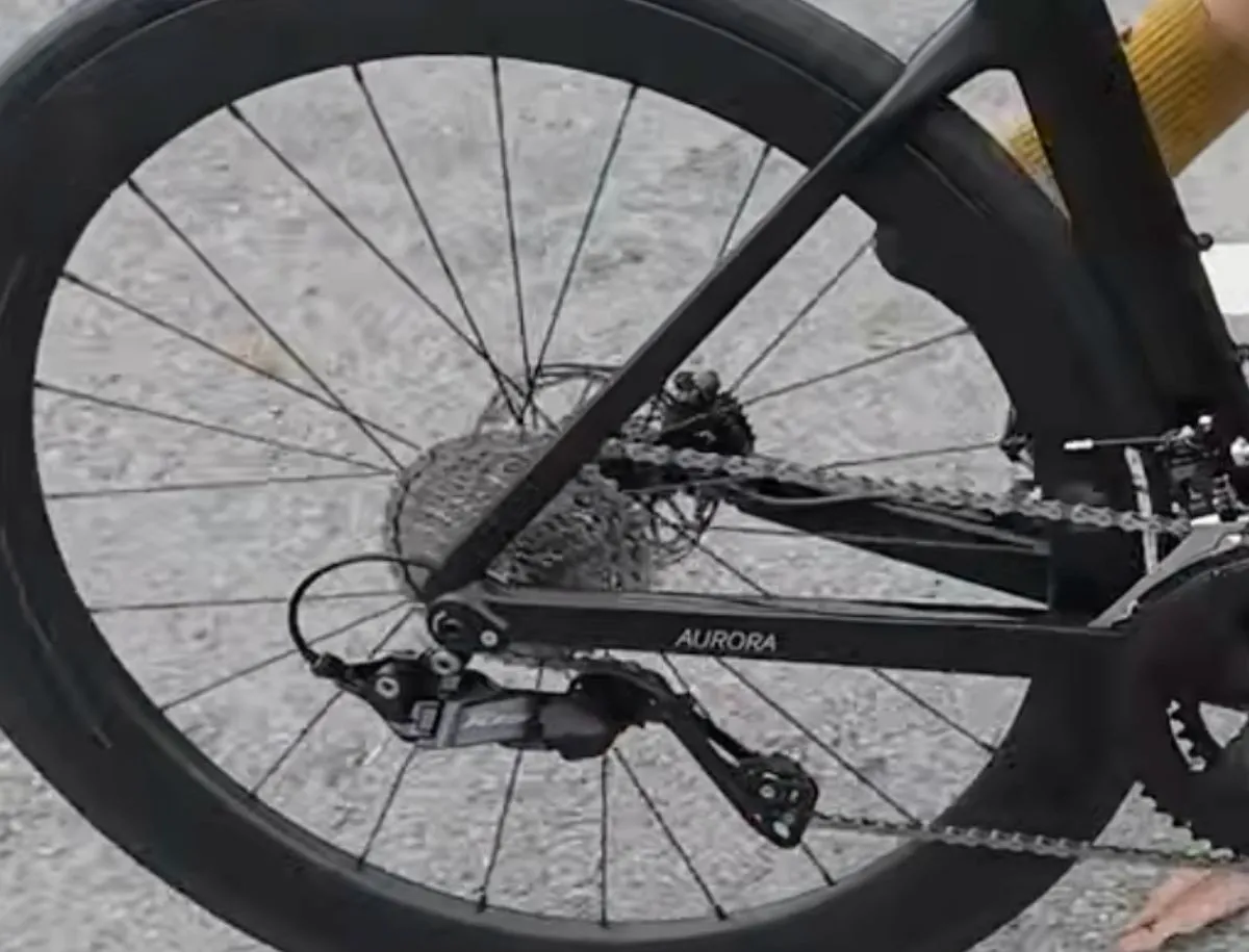 Close-up of Shimano 105 R7120 rear derailleur and cassette on SAVA A7L Pro carbon road bike during a ride, showing Aurora chainstay label and deep-section rear carbon wheel in the background