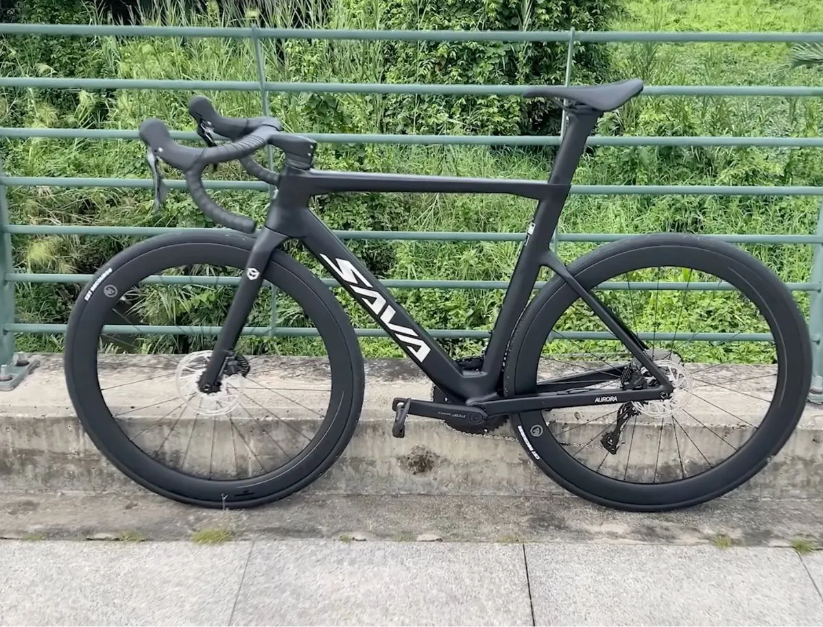 Full side profile of matte black SAVA Aurora A7L Pro carbon road bike leaning against a green railing, showing deep-section carbon wheelset, integrated handlebar, dropped seatstay design, and Shimano disc brakes