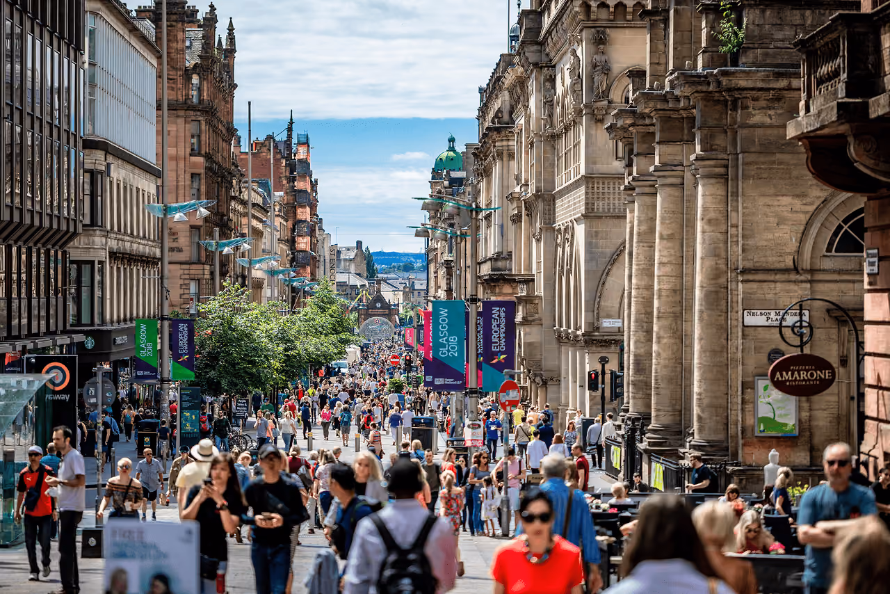 Buchanan Street pedestrian zone packed with summer shoppers flanked by grand sandstone buildings and Glasgow 2018 European Championships banners