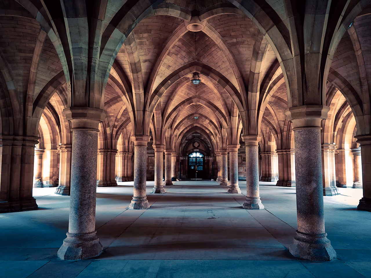 The vaulted Gothic sandstone cloisters of Glasgow University with rows of columns receding toward a stained glass window
