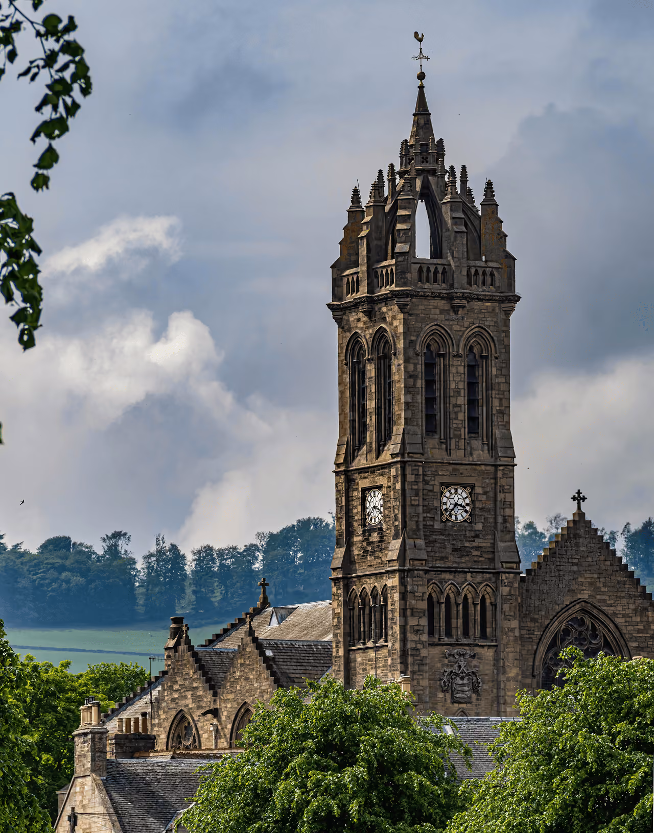 The ornate Gothic clock tower of a Borders church rising above rooftops and summer trees against a dramatic sky