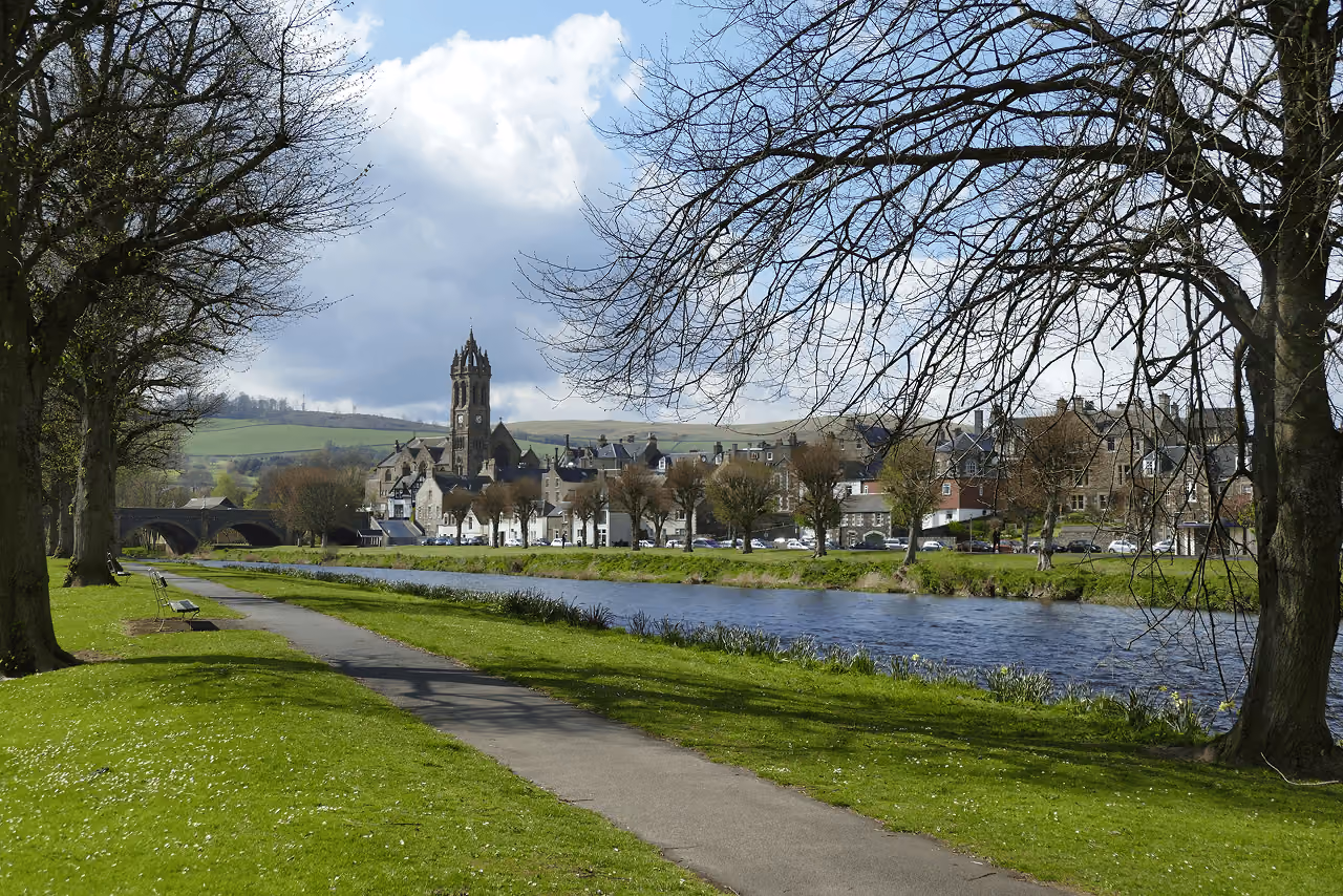 A riverside walkway in Peebles with bare spring trees, a stone bridge and the town's church tower visible beyond