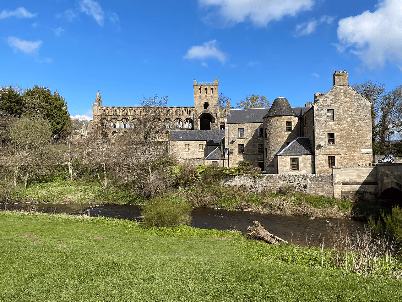 Jedburgh Abbey ruins towering over a stone building beside the River Jed on a bright spring day