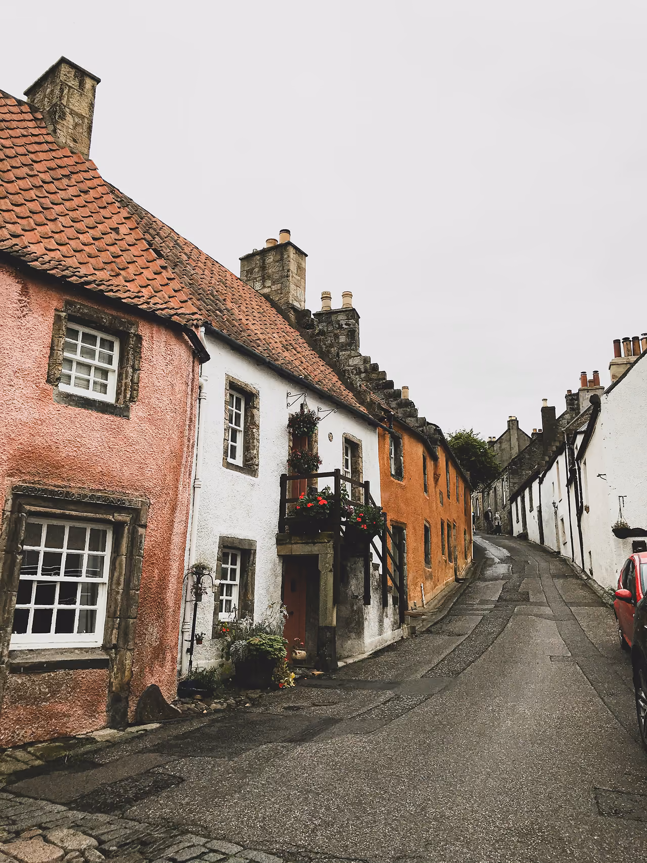 A narrow lane in Culross lined with pink, white and terracotta historic cottages with tiled roofs on a quiet grey day