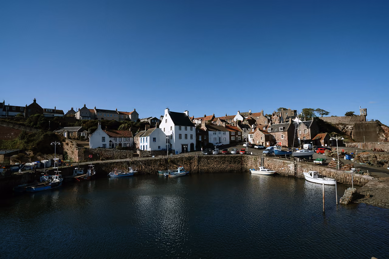 Crail's small stone harbour with fishing boats moored below whitewashed and sandstone buildings stacked up the hillside on a clear sunny day