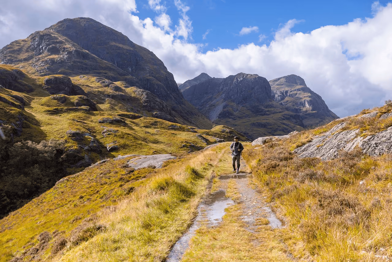 A lone hiker on a rocky path through Glencoe with the Three Sisters rising dramatically on both sides under a bright blue sky