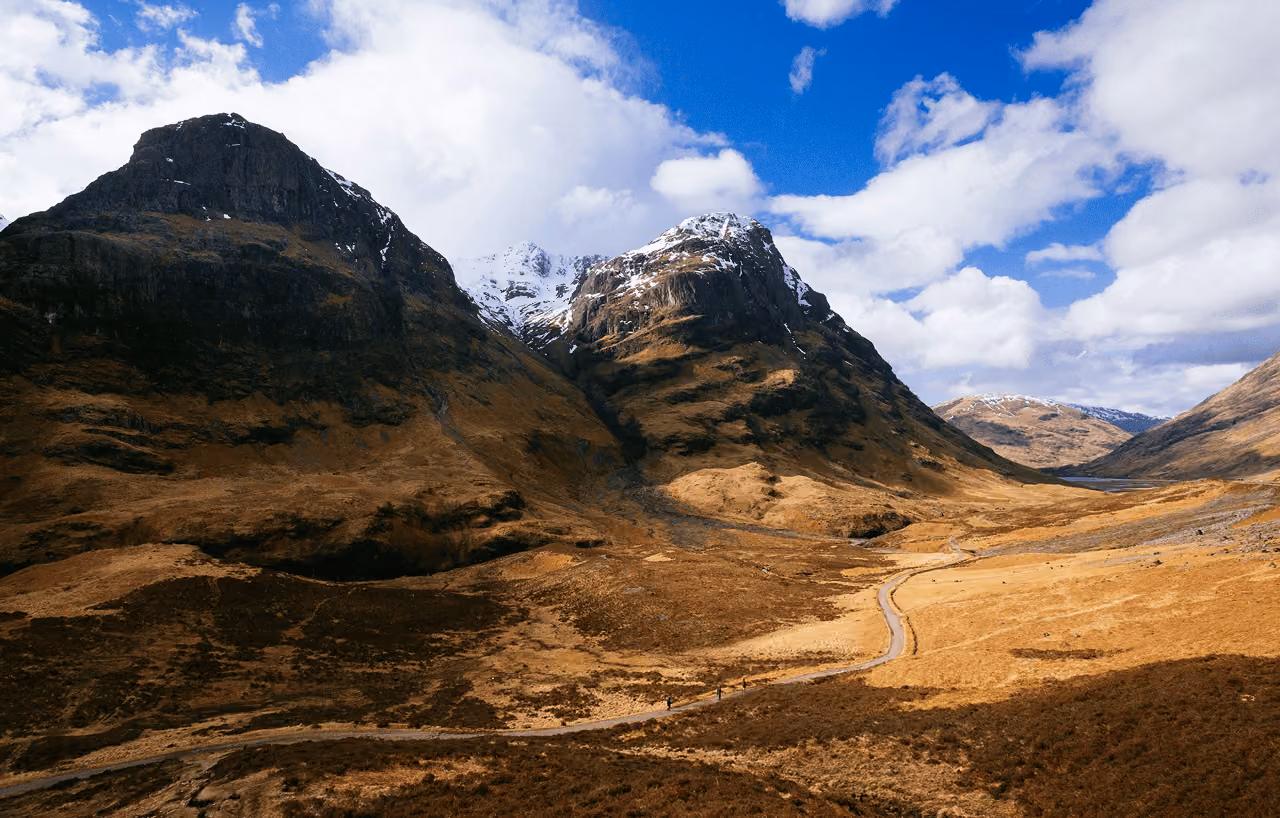 The Three Sisters of Glencoe rising above a golden sunlit valley floor with snow on the peaks and the A82 road below