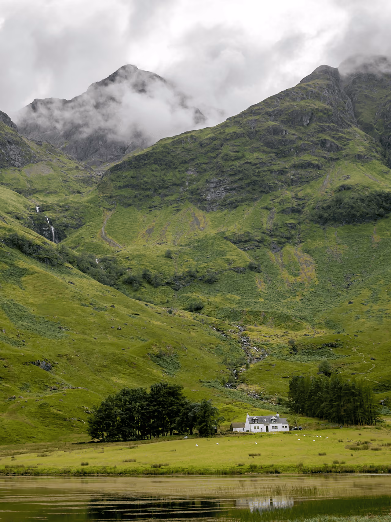 A white cottage beside Loch Achtriochtan dwarfed by towering green mist-shrouded mountains with a waterfall cascading in the background