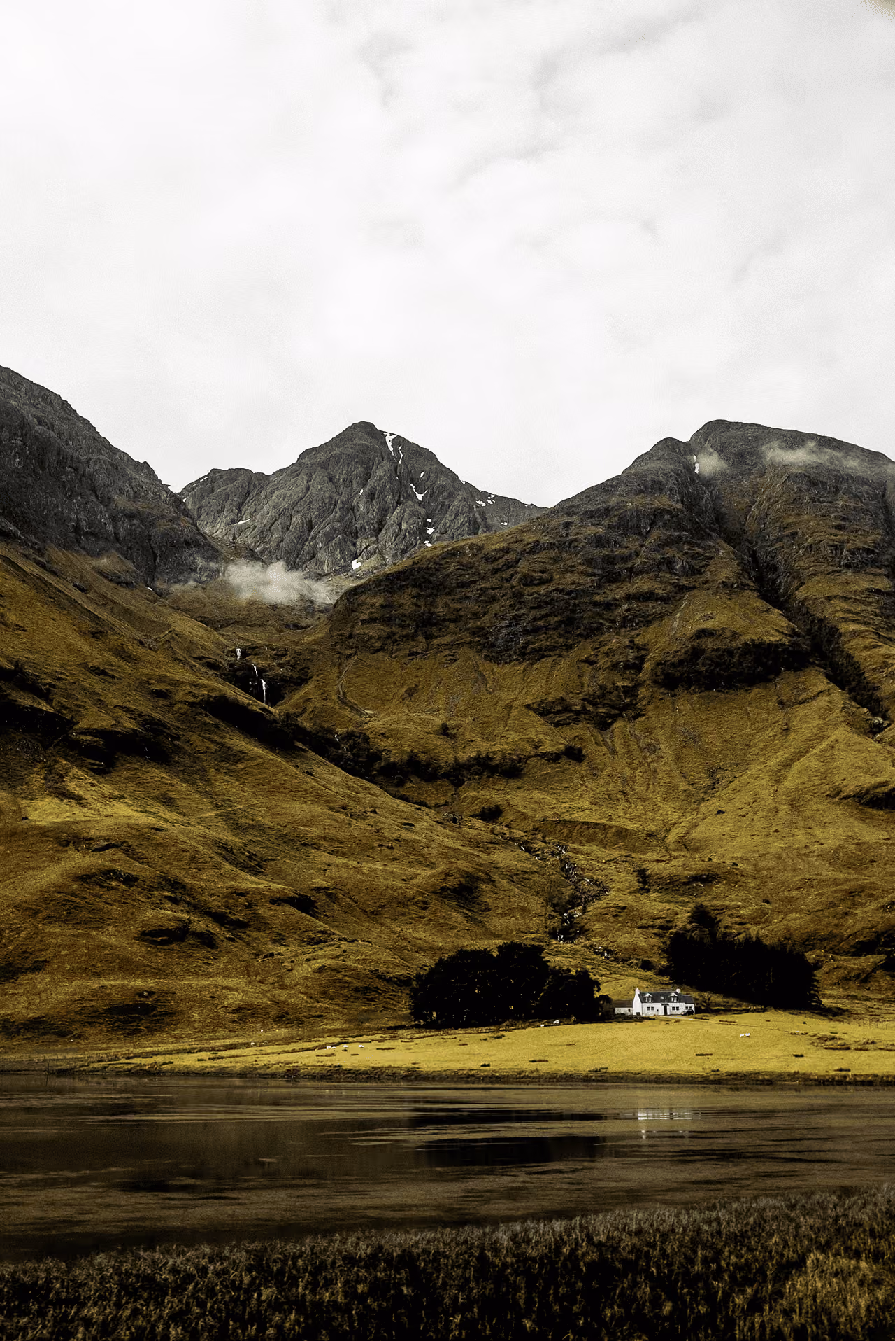 The same lone white cottage below the stark rocky Three Sisters peaks with snow patches and low cloud in moody winter light