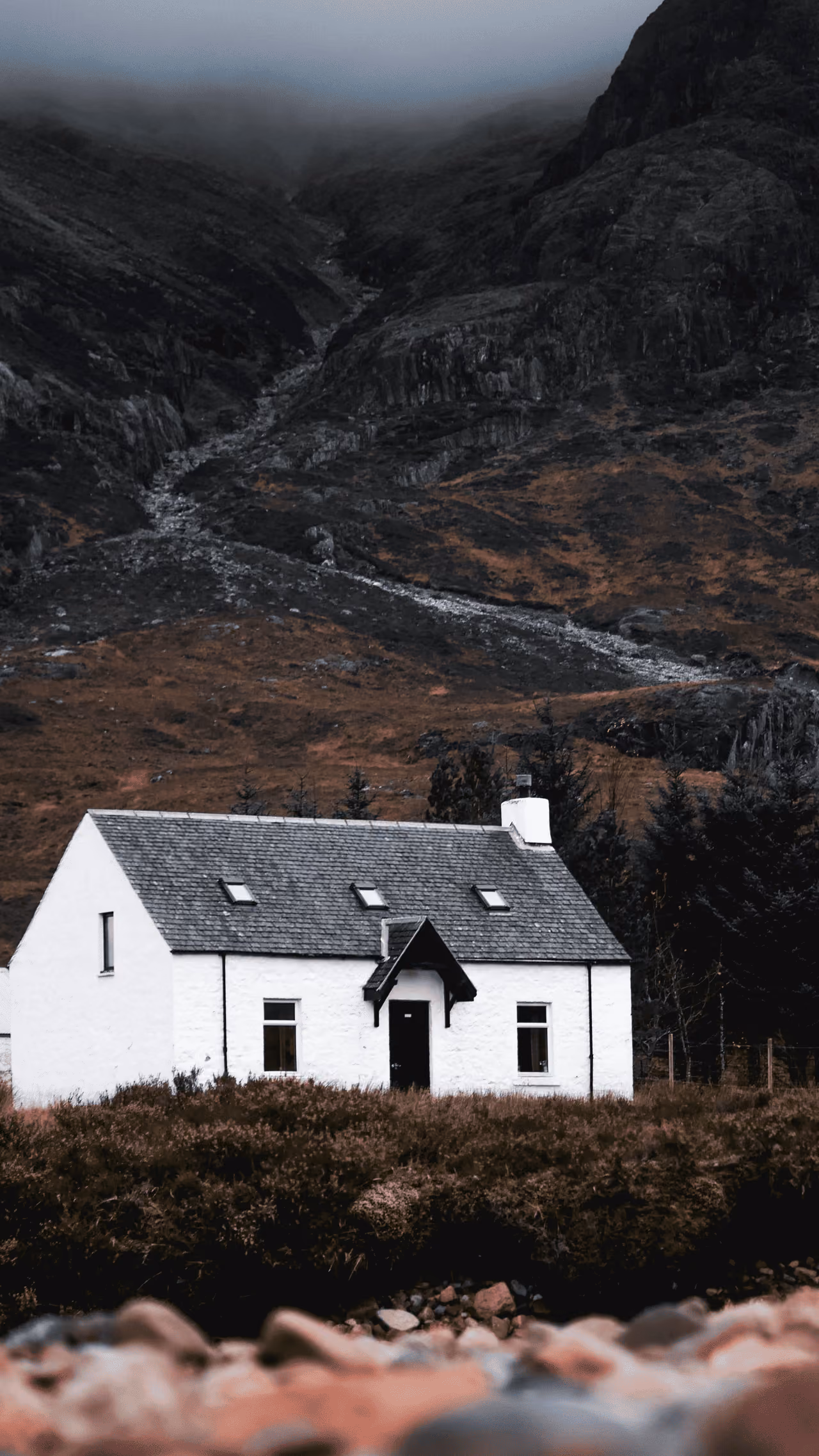 A whitewashed Highland cottage in Glencoe framed by dark rocky mountains and autumn moorland under a grey overcast sky
