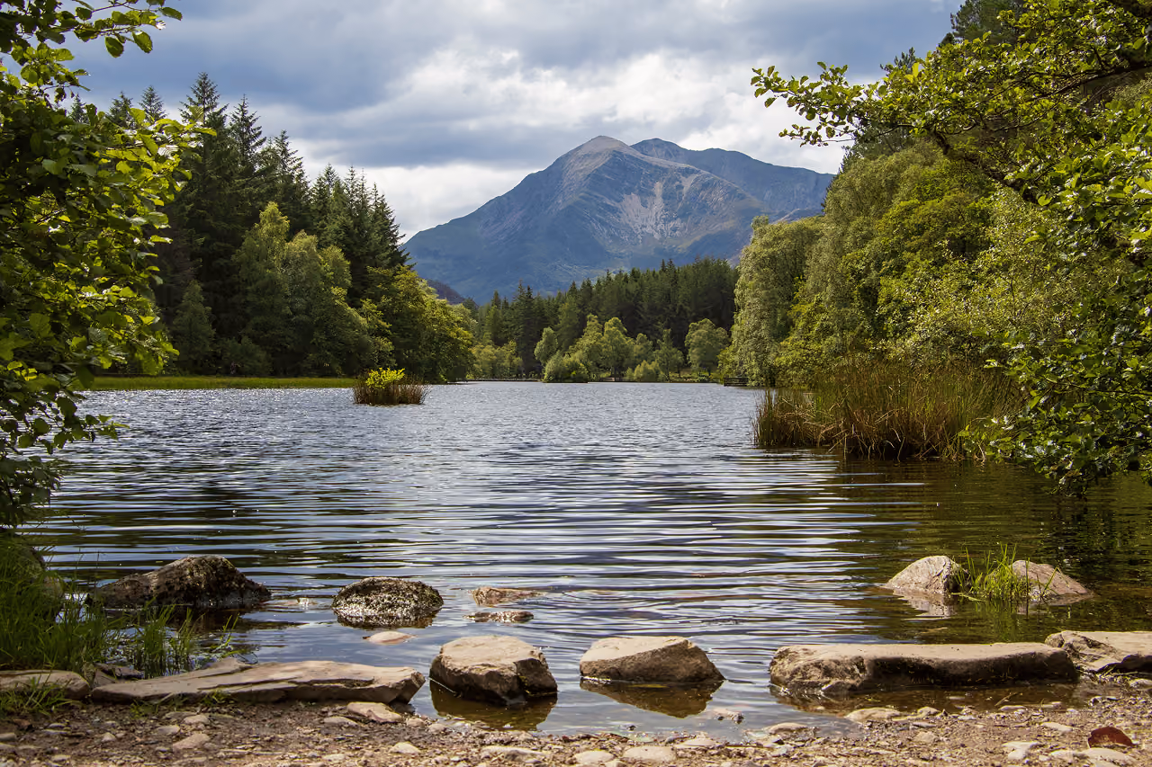 Glencoe Lochan's rocky shoreline framed by summer trees with a towering mountain peak rising behind under a cloudy sky