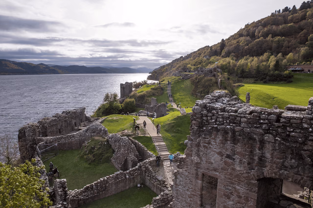 Visitors exploring the extensive stone ruins of Urquhart Castle perched above Loch Ness with forested hillsides stretching into the distance