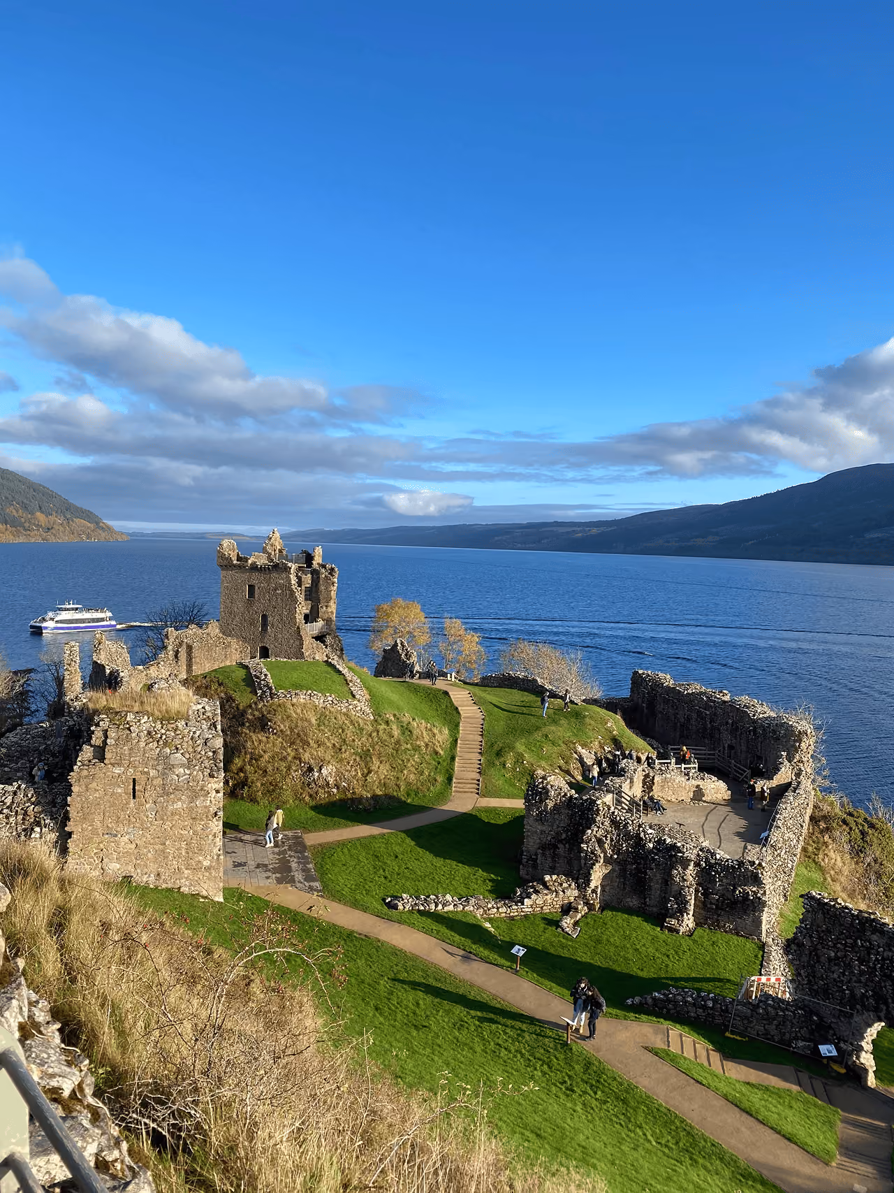 Urquhart Castle ruins in bright autumn sunshine with a white tour boat passing on the sparkling blue waters of Loch Ness
