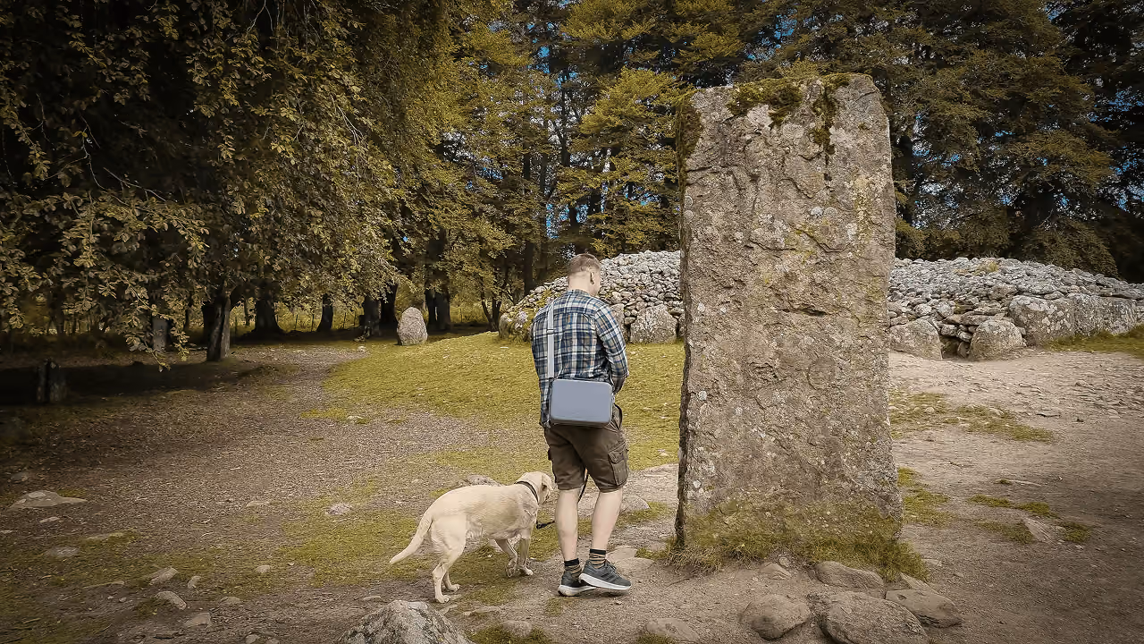 A visitor with a yellow labrador beside a tall standing stone at Clava Cairns with a stone cairn and trees behind