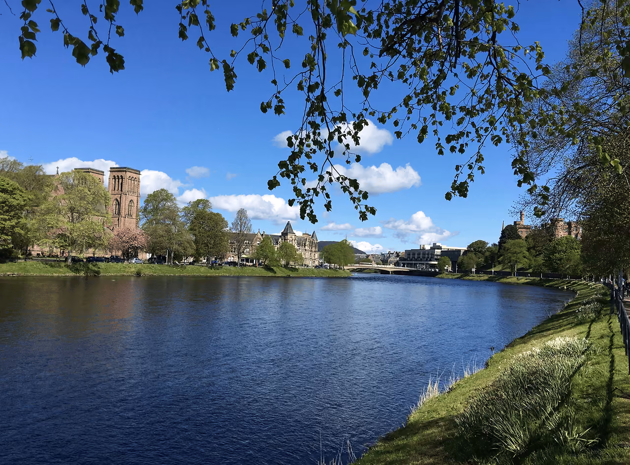 Inverness Cathedral's twin towers framed by overhanging spring branches beside the sparkling River Ness on a sunny blue-sky day