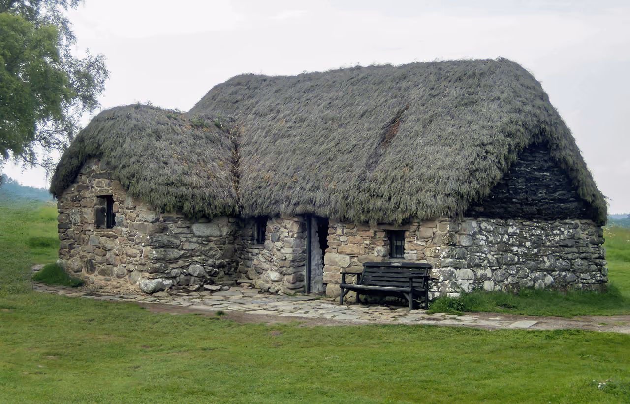 Leanach Cottage at Culloden Battlefield, a low traditional thatched stone building with a wooden bench beside the entrance on a grey overcast day