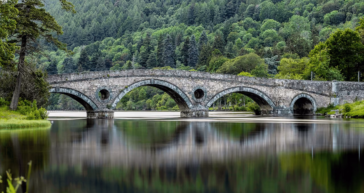 Kenmore Bridge, a graceful five-arched stone bridge reflected in the still waters of the River Tay surrounded by dense green forest