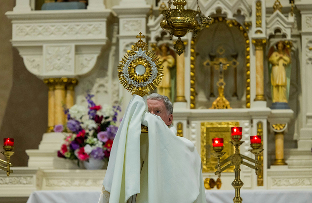 Priest holding a gold monstrance during a religious ceremony inside a church altar area.