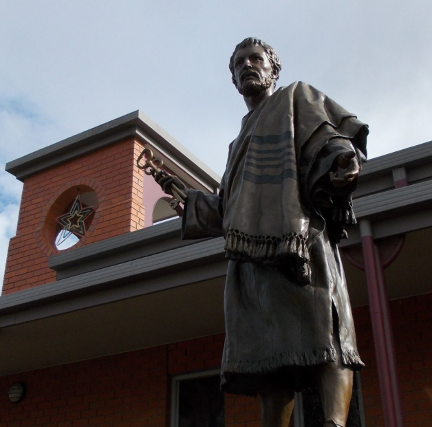 Bronze statue of a bearded man holding large keys, standing in front of a brick building.