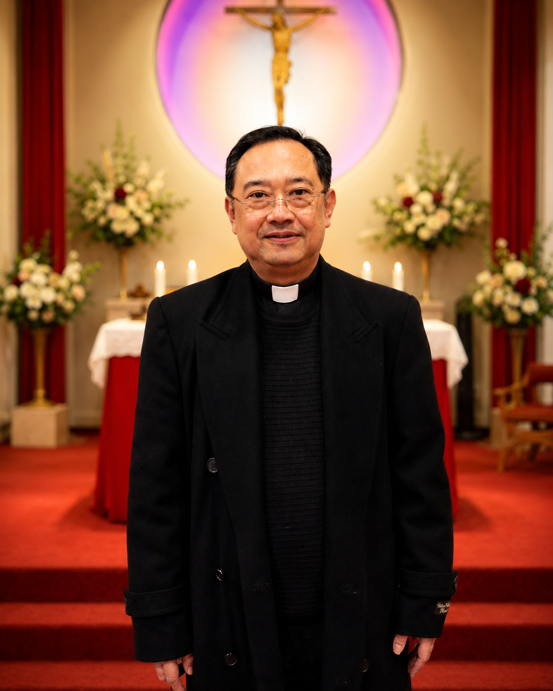Man in white clerical robe and glasses in front of a statue and candle in a wood-paneled room.