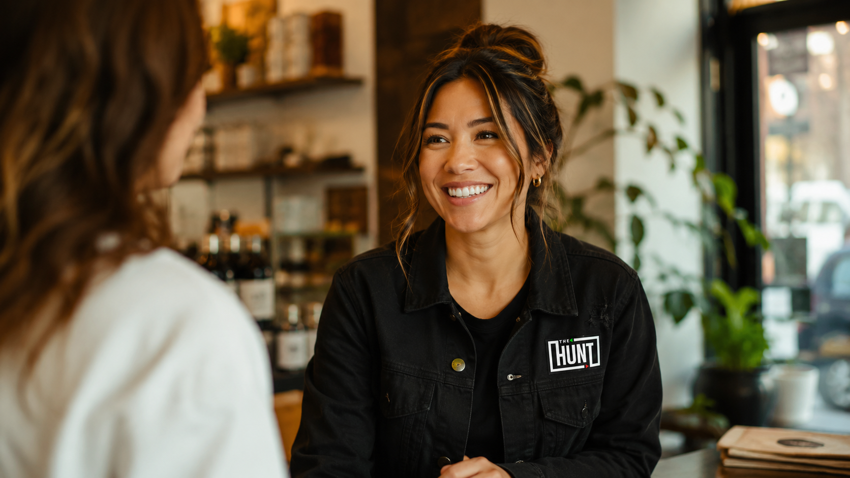 Smiling woman in a black jacket with The Hunt logo conversing with another person indoors.
