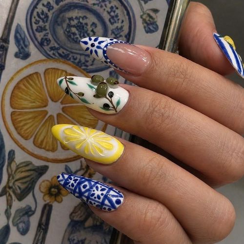 Hand with almond-shaped nails painted with blue patterns, lemons, and olives on white background.