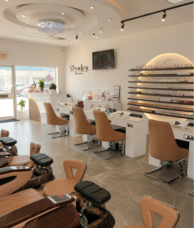 Modern nail salon interior with tan chairs, manicure stations, a nail polish display, and a chandelier.
