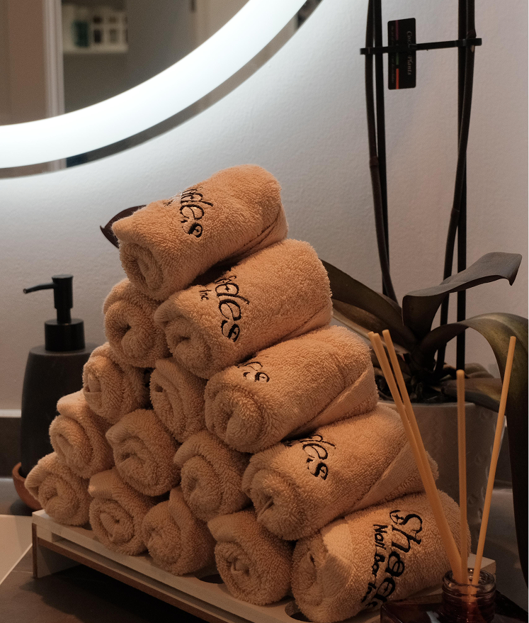 Stack of rolled beige towels on a tray next to a plant and scent sticks in a spa setting.