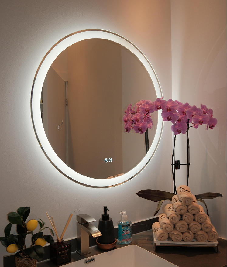 Circular backlit mirror above sink with orchid plant and stacked rolled towels on counter.