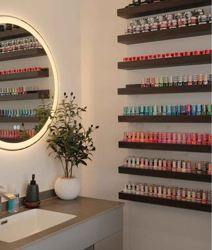 Bathroom countertop with a sink, plant, soap dispenser, and shelves of colorful nail polish bottles.