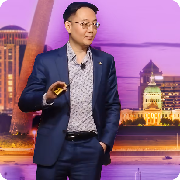 Man in glasses and a suit holding a clicker stands before a cityscape with illuminated buildings at dusk.