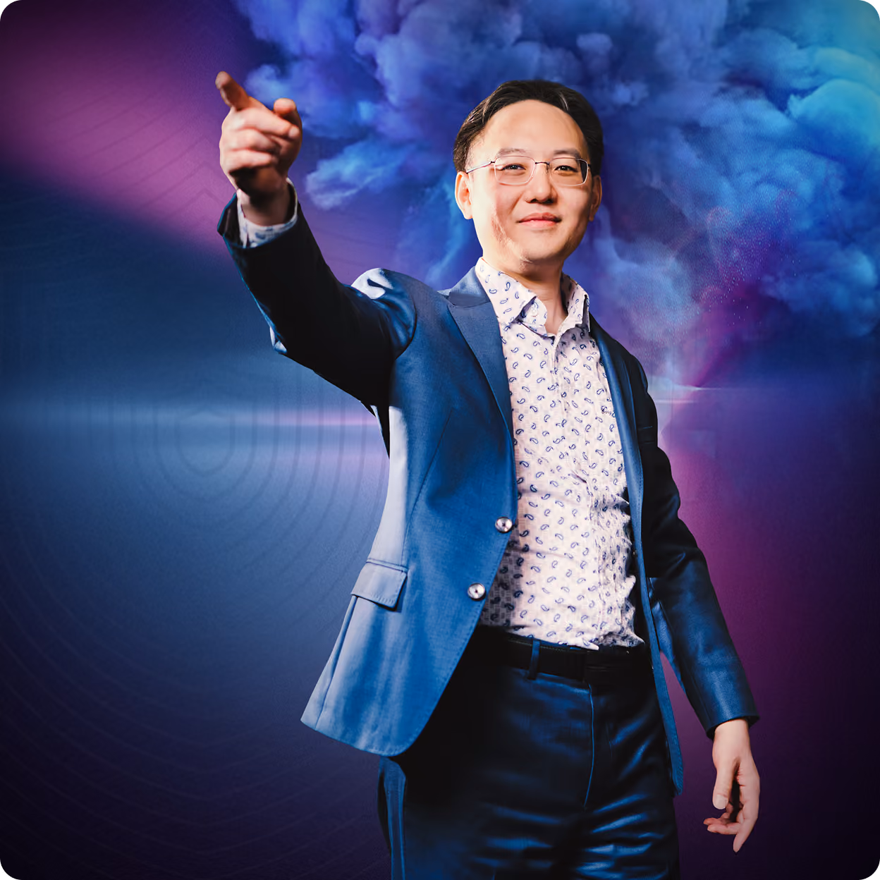 Man in blue suit and patterned shirt pointing forward against a blue and purple clouded background.