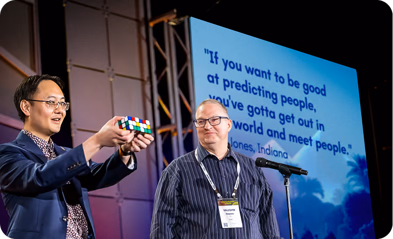 Two men at a conference, one holding a Rubik's cube, the other by a microphone.